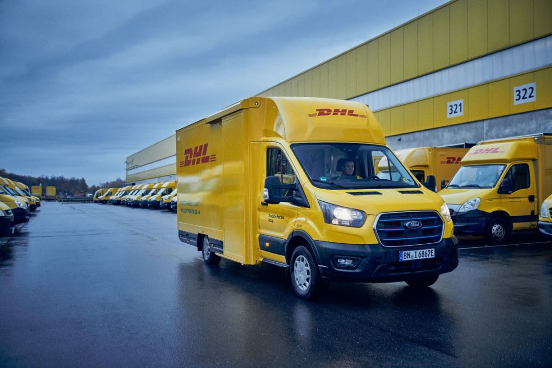 A BRIGHT YELLOW STRAIGHT SIDED DELIVERY VAN BEARING THE DHL LOGO DRIVES DOWN THE STREET
